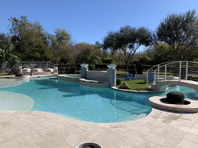Freeform Sugar Land pool with a curved footbridge, raised spa, and travertine deck maintained by Lanterra Pools