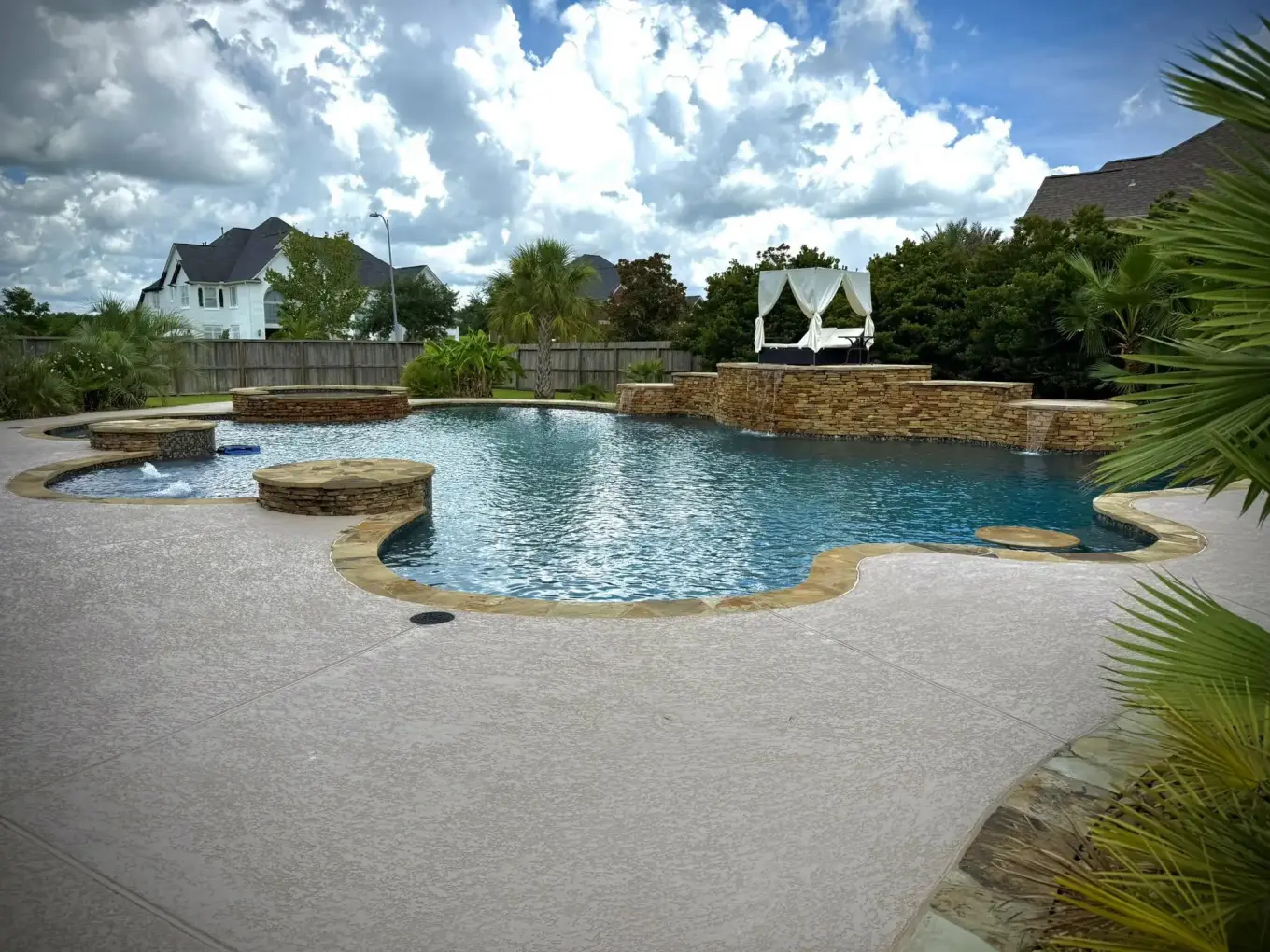 Large freeform pool with a stacked stone waterfall wall, raised spa, and cabana under summer clouds